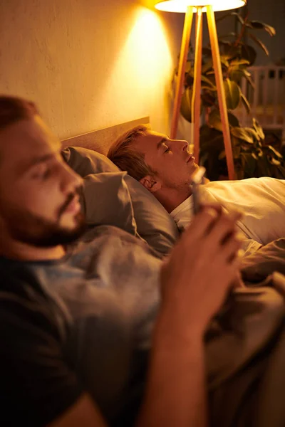 Unfaithful gay man browsing date app on mobile phone near sleeping boyfriend at night in bedroom — Stock Photo