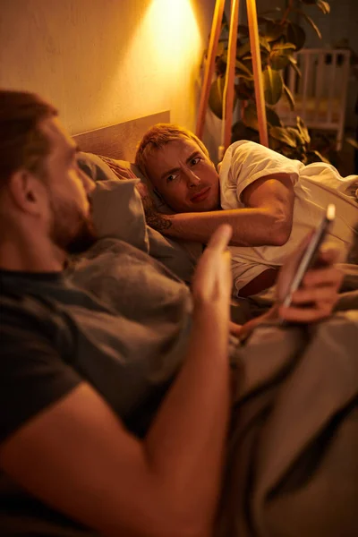 Disloyal gay man browsing date app on smartphone near discouraged boyfriend at night in bedroom — Stock Photo