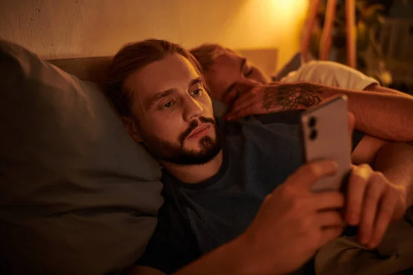 Disloyal bearded gay man browsing date app on smartphone near sleeping boyfriend at night in bedroom — Stock Photo