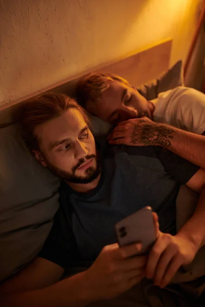 Unfaithful bearded gay man chatting on smartphone near sleeping boyfriend at night in bedroom — Stock Photo