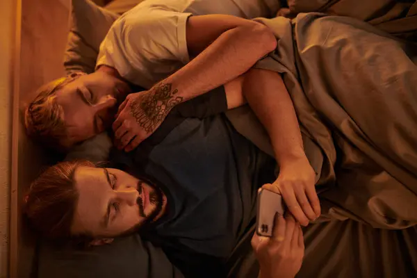 Top view of unfaithful bearded gay man chatting on smartphone near sleeping boyfriend at night — Stock Photo