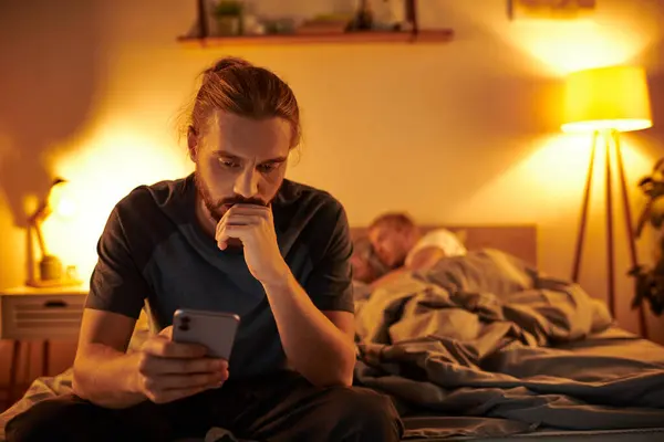 Лояльний бородатий гей-чоловік переглядає Інтернет на смартфоні біля партнера, який спить вночі в спальні — стокове фото