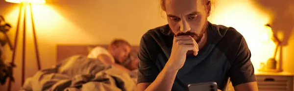 Disloyal gay man browsing internet on smartphone near partner sleeping at night in bedroom, banner — Stock Photo