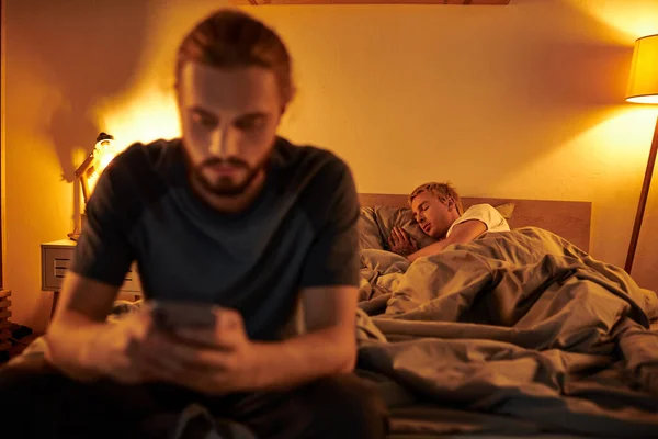 Disloyal bearded gay man browsing internet on smartphone near partner sleeping at night in bedroom — Stock Photo
