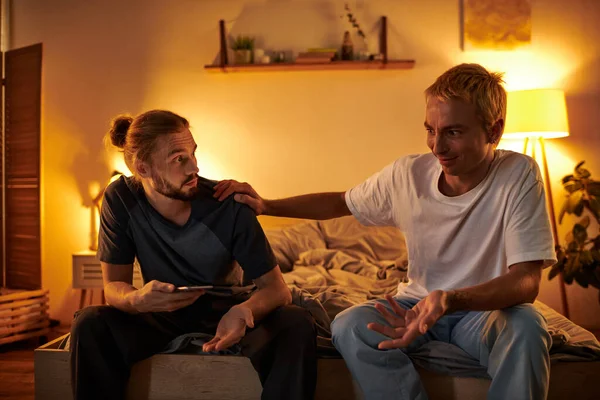 Discouraged gay man quarreling with boyfriend sitting with smartphone at night in bedroom, cheating — Stock Photo