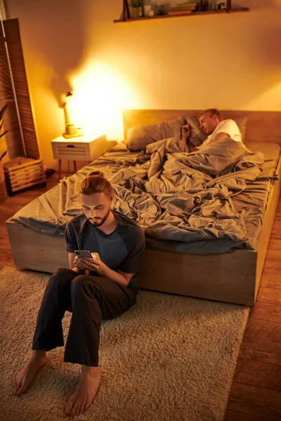 Unfaithful bearded gay man messaging on mobile phone near sleeping boyfriend at night in bedroom — Stock Photo