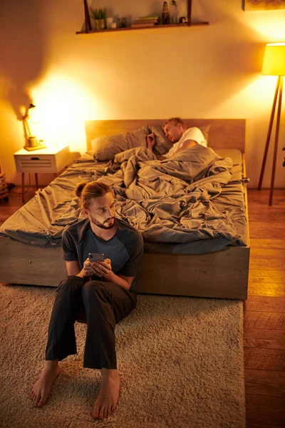 Disloyal bearded gay man browsing date app near sleeping boyfriend at night in bedroom, cheating — Stock Photo