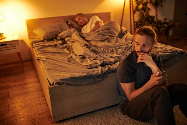 Disloyal gay man browsing date app on smartphone near sleeping boyfriend at night in bedroom — Stock Photo