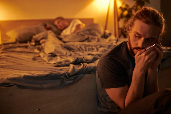 Disloyal bearded gay man talking on mobile phone near sleeping boyfriend at night in bedroom — Stock Photo