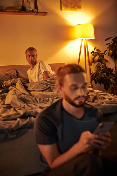 Discouraged gay man looking at boyfriend messaging on mobile phone in bedroom at night, cheating — Stock Photo