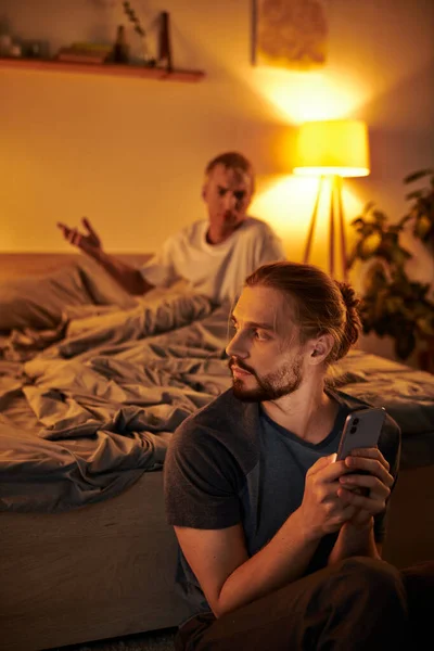 Displeased gay man quarreling with boyfriend messaging on smartphone at night in bedroom, trouble — Stock Photo