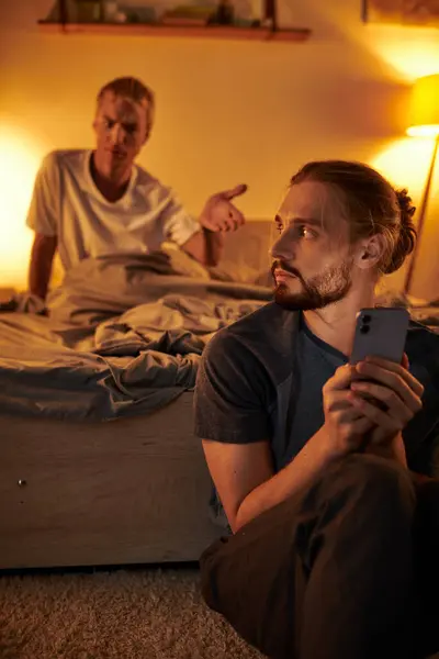 Displeased gay man quarreling with boyfriend messaging on smartphone at night in bedroom, trouble — Stock Photo