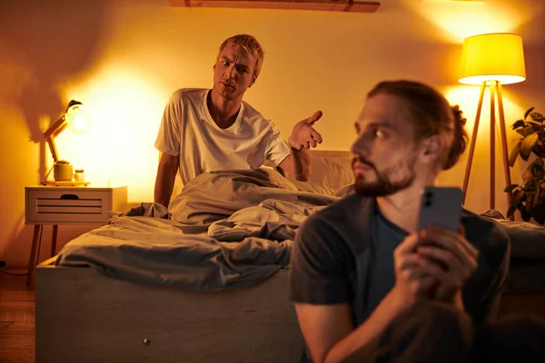 Displeased gay man quarreling with boyfriend messaging on smartphone at night in bedroom, trouble — Stock Photo