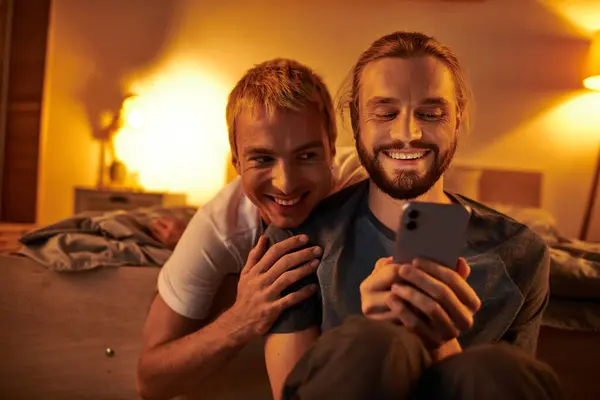 Joyeuse barbu gay homme navigation internet sur téléphone mobile près de sourire petit ami dans la chambre à coucher la nuit — Photo de stock