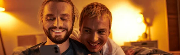 Alegre gay hombre navegación internet en móvil teléfono cerca sonriente novio en habitación en la noche, banner - foto de stock