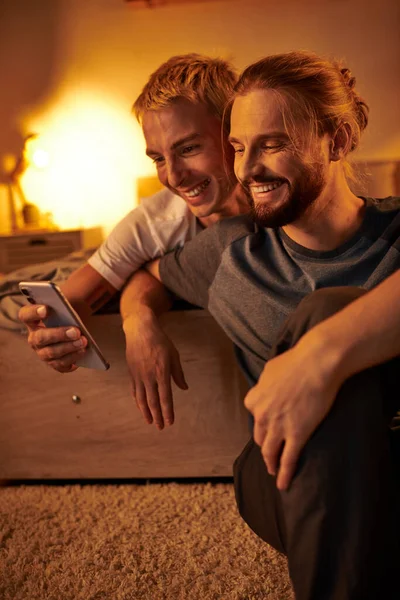 Joyful bearded gay man browsing internet on mobile phone near smiling boyfriend in bedroom at night — Stock Photo