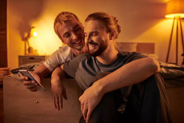 Joyful bearded gay man browsing internet on mobile phone near smiling boyfriend in bedroom at night — Stock Photo