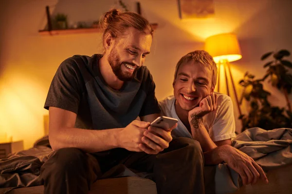 Happy bearded gay man browsing internet on mobile phone near smiling boyfriend in bedroom at night — Stock Photo