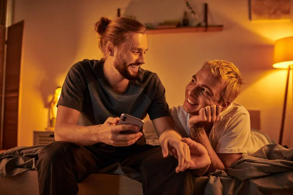 Happy bearded gay man browsing internet on mobile phone near smiling boyfriend in bedroom at night — Stock Photo