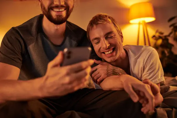 Smiling bearded gay man browsing internet on mobile phone near happy boyfriend in bedroom at night — Stock Photo