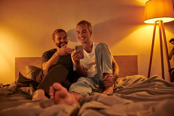 Smiling gay man browsing internet on mobile phone near happy bearded boyfriend in bedroom at night — Stock Photo