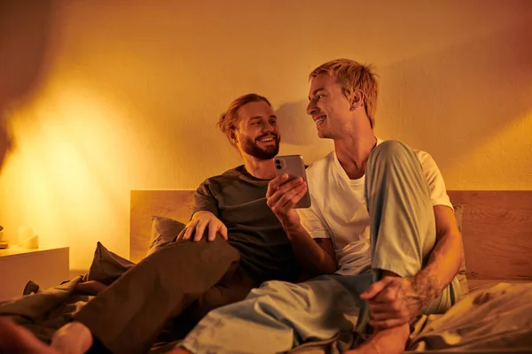 Smiling gay man using mobile phone near happy bearded boyfriend in bedroom at night, tranquility — Stock Photo