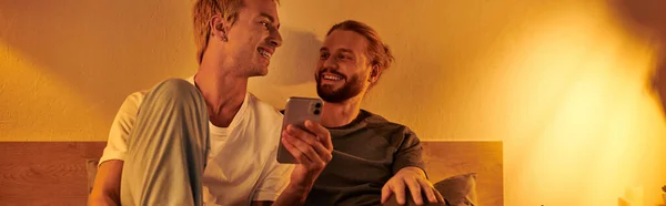 Smiling gay man using mobile phone near happy bearded boyfriend in bedroom at night, banner — Stock Photo