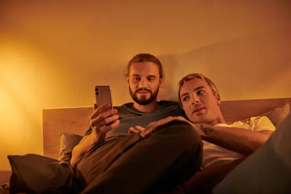 Happy bearded gay man browsing social media on smartphone near smiling boyfriend in bedroom at night — Stock Photo