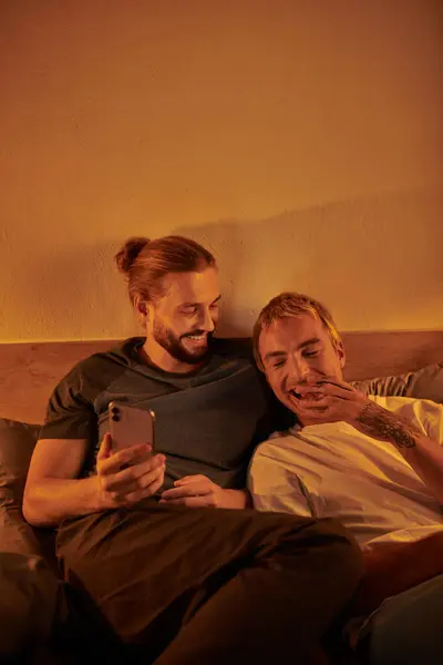 Bearded gay man browsing social media on smartphone near laughing boyfriend in bedroom at night — Stock Photo