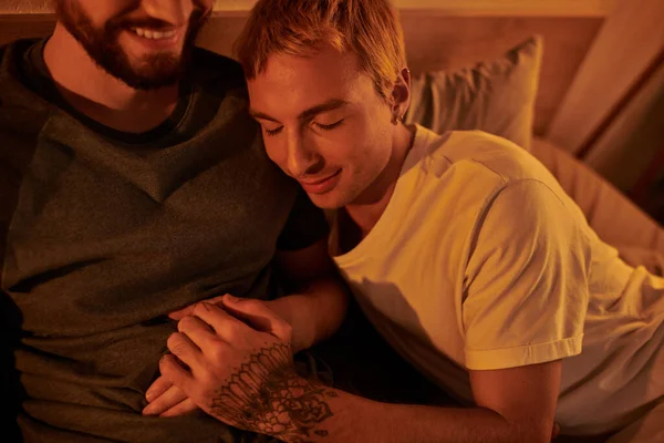 Tatuado gay hombre sonriendo con cerrado ojos cerca feliz barbudo novio en la noche en dormitorio gay fecha - foto de stock
