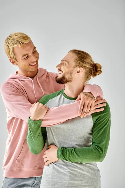 Fröhliches homosexuelles Paar in stilvoller Freizeitkleidung, das sich umarmt und einander auf grauem Hintergrund ansieht — Stockfoto