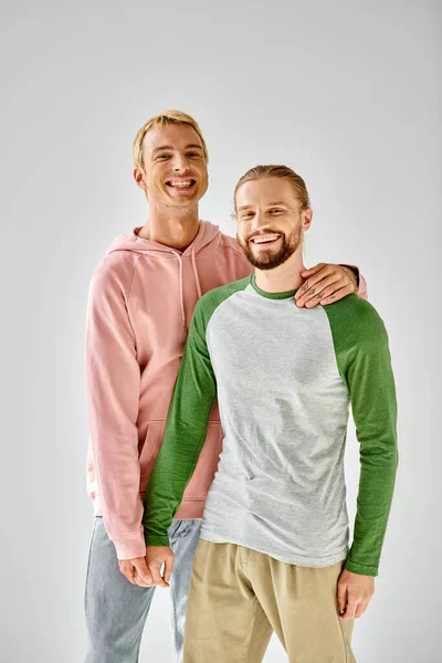 Joyful gay couple in stylish casual attire standing and looking at camera on grey backdrop — Stock Photo