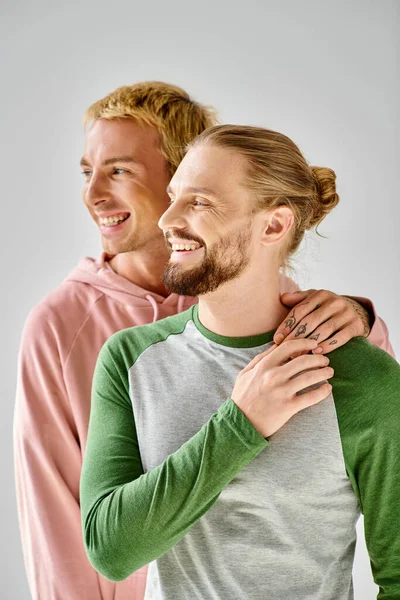 Joyful gay couple in stylish casual attire standing and looking away on grey backdrop in studio — Stock Photo