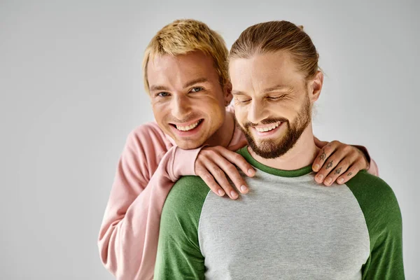 Feliz gay hombre en casual traje abrazando hombros de barbudo novio y mirando la cámara en gris - foto de stock