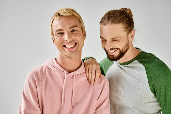 Alegre gay hombre en casual atuendo sonriendo en cámara cerca barbudo novio en gris estudio telón de fondo - foto de stock