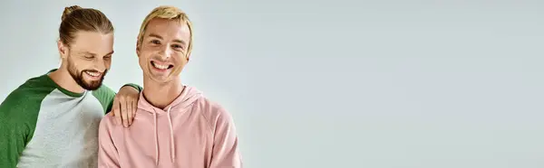 Fröhlicher schwuler Mann in legerer Kleidung, der in die Kamera lächelt, neben bärtigem Freund auf grauem Hintergrund, Banner — Stockfoto