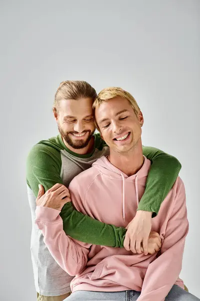 Alegre barbudo gay hombre abrazando elegante novio sonriendo con cerrado ojos en gris telón de fondo - foto de stock