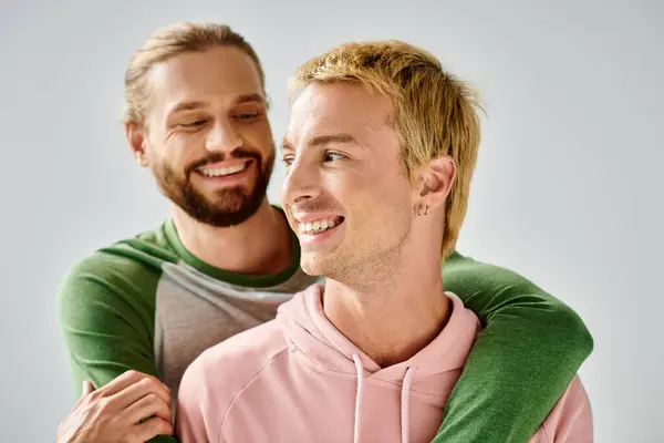 Fröhlicher bärtiger schwuler Mann umarmt stilvollen Freund lächelnd und schaut vor grauem Hintergrund weg — Stockfoto