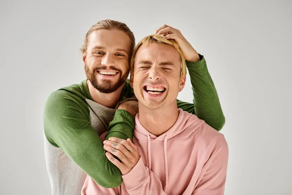 Cheerful bearded gay man embracing trendy boyfriend while having fun on grey backdrop in studio — Stock Photo