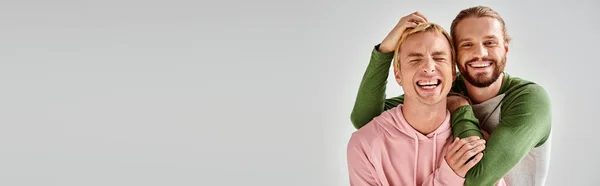 Cheerful bearded gay man embracing trendy boyfriend while having fun on grey backdrop, banner — Stock Photo