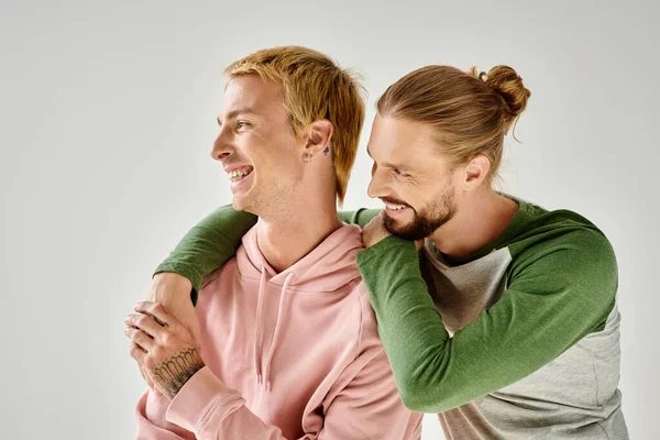 Fröhliches und stilvolles schwules Paar, das sich umarmt und wegsieht, während es vor grauem Hintergrund steht — Stockfoto