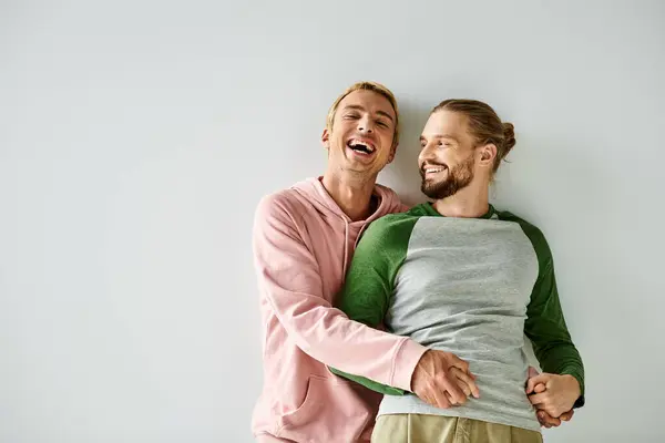 Excited gay man holding hands of bearded boyfriend and laughing on grey backdrop, same-sex couple — Stock Photo