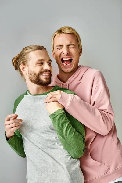 Überglücklicher und trendiger schwuler Mann lacht mit geschlossenen Augen in der Nähe eines bärtigen Freundes vor grauem Hintergrund — Stockfoto