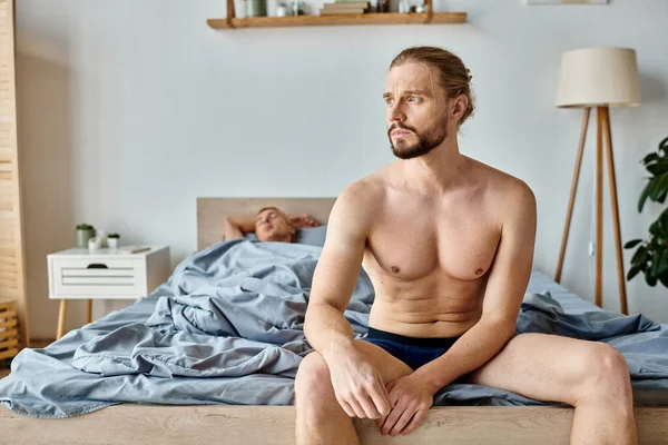 Sconvolto uomo barbuto in mutande seduto e guardando lontano vicino partner amore dormire in camera da letto — Foto stock