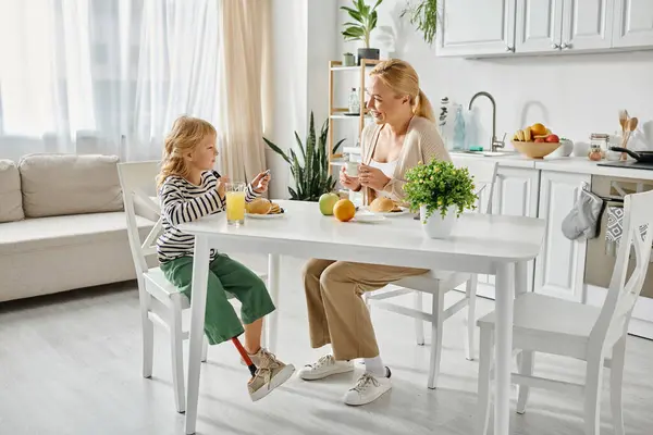 Preschooler girl with prosthesis leg having breakfast with happy blonde mother in modern kitchen — Stock Photo