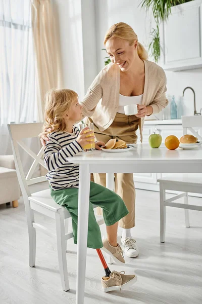 Menina pré-escolar com perna prótese tomando café da manhã com mãe loira feliz na cozinha moderna — Fotografia de Stock