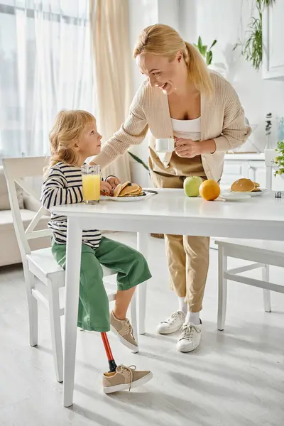 Happy mother standing near daughter with prosthetic leg having breakfast in kitchen, inclusion — Stock Photo