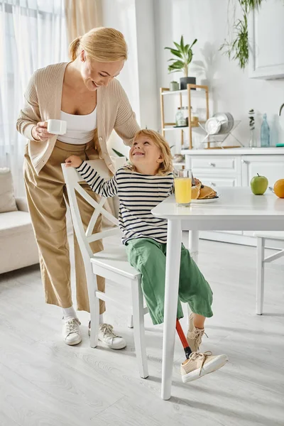 Glückliche Mutter, die in der Nähe von niedlichen Mädchen mit Beinprothese beim Frühstück in der Küche steht, Inklusion — Stockfoto