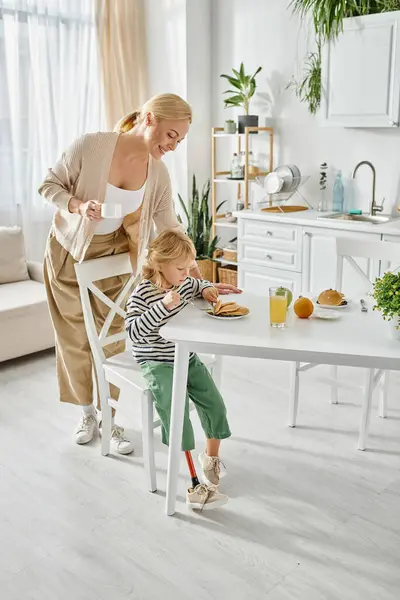 Donna felice in piedi vicino a figlia con gamba protesica che fa colazione in cucina, inclusione — Foto stock