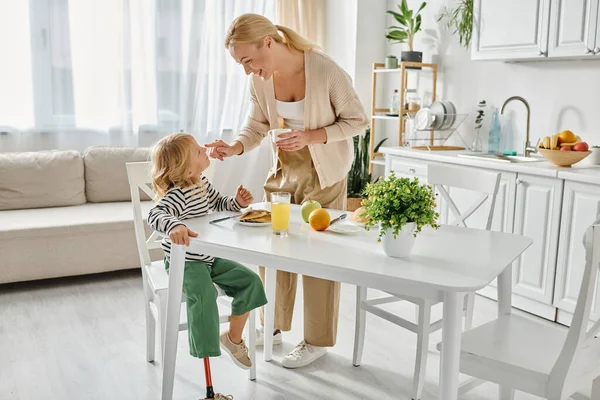 Happy mother standing near daughter with prosthetic leg having breakfast in kitchen, disability — Stock Photo